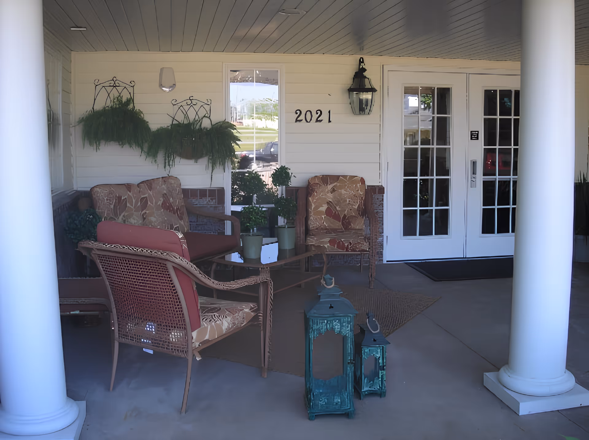 Covered porch area with white columns, cushioned wicker chairs and a loveseat arranged around a glass coffee table with potted plants. Two decorative green lanterns are on the floor in front. The wall behind has hanging ferns, a window, a black lantern light fixture, and the number 2021. Double glass doors lead inside.