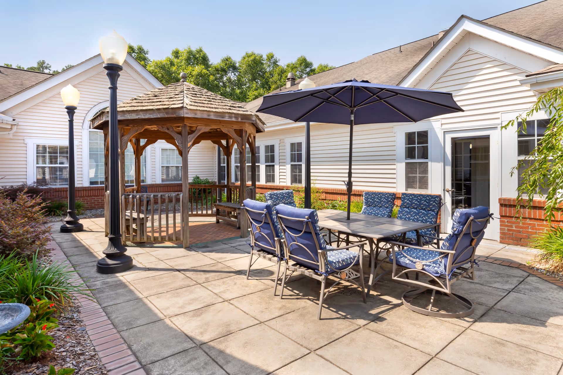 Outdoor patio area at Brookdale Cary featuring a wooden gazebo, a table with six cushioned chairs and a large umbrella, surrounded by a paved floor and garden plants, with white siding buildings in the background under a clear sky.