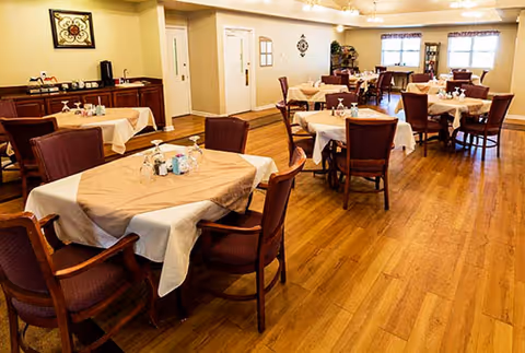 A dining room with several tables covered with beige tablecloths, each surrounded by wooden chairs with cushioned seats. The room has wooden flooring, light-colored walls, and windows letting in natural light. There is a sideboard with coffee and tea supplies along one wall.
