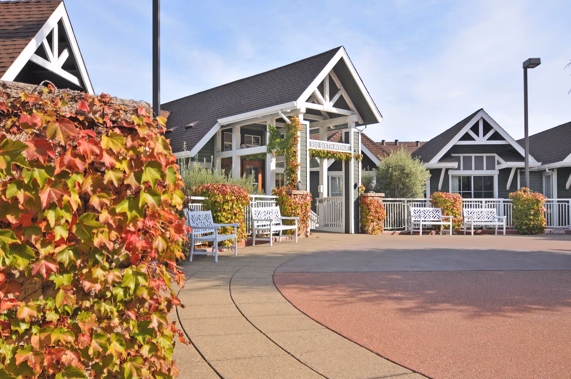 Exterior view of a senior living facility with a paved walkway, white benches, and buildings featuring dark roofs and white trim. There are plants with red and green leaves growing on the walls and around the entrance area. The sky is clear and blue.