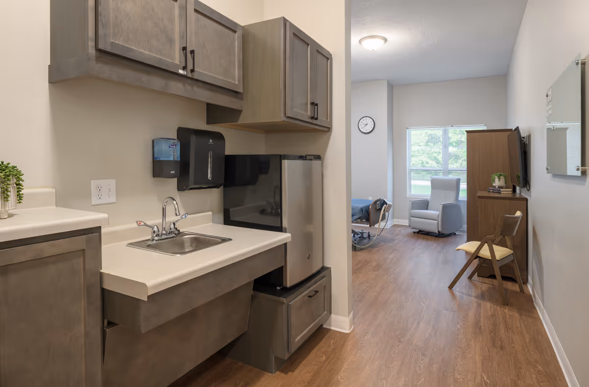 Interior view of a senior living facility room showing a small kitchenette with a sink, cabinets, and a mini refrigerator. The room extends into a living area with a recliner chair, a wooden cabinet, a wall-mounted TV, a chair, and a window letting in natural light. The floor is wood, and the walls are painted light gray.