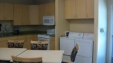 Compact kitchen with light wood cabinets, appliances, and a white dining table surrounded by chairs.