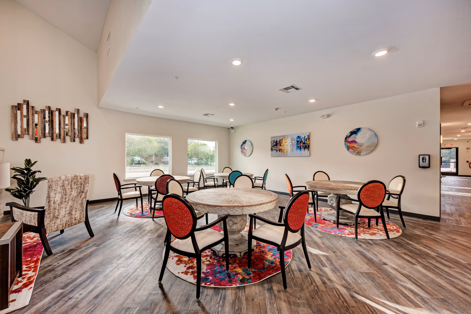 A bright and spacious common area with round wooden tables and chairs featuring red and beige upholstery. The floor has a wood finish with colorful circular rugs under each table. The walls are decorated with modern abstract art, and large windows allow natural light to fill the room. There is a comfortable armchair and a potted plant in one corner.