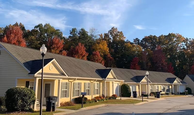 Exterior view of a single-story residential building with multiple units, each having a front porch and windows with shutters. The building is light yellow with a dark gray roof. There are street lamps and trash bins along the sidewalk, and colorful autumn trees in the background.