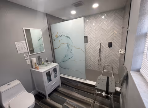Accessible bathroom with shower stall, toilet, vanity, and fold-up chair in a senior facility.