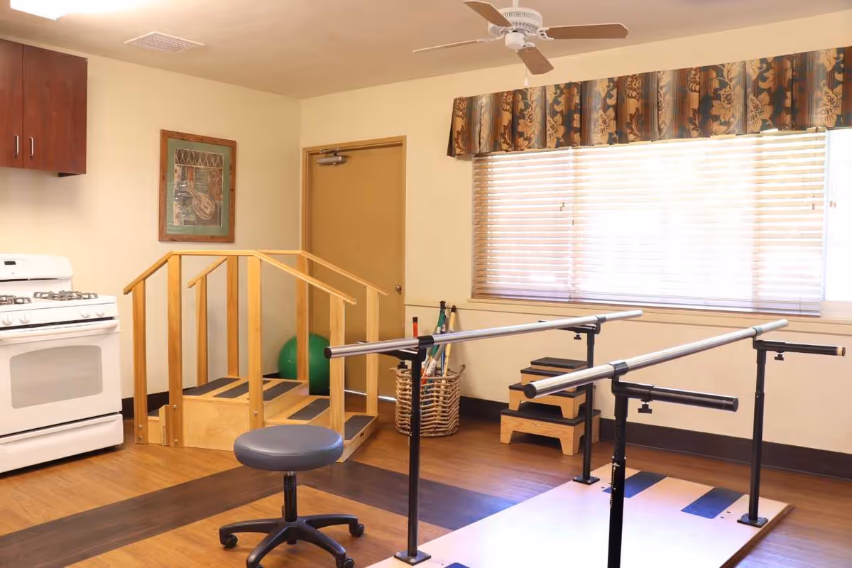 A rehabilitation room with parallel bars for walking exercises, a small wooden staircase with handrails, a green exercise ball, a stool on wheels, a white stove, wooden cabinets, a window with blinds and floral valance, and a ceiling fan.