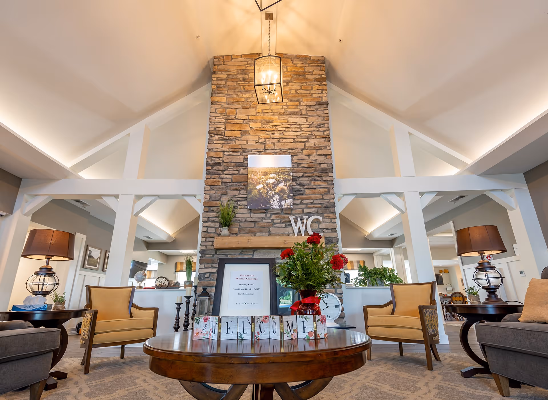 A cozy living room area with a high vaulted ceiling and a stone fireplace in the center. The fireplace has a framed picture and decorative letters 'WC' on the mantel. In front of the fireplace is a round wooden table with a flower arrangement and blocks spelling 'WELCOME'. The room features comfortable seating with armchairs and sofas, side tables with lamps, and decorative plants. The space is warmly lit and inviting.