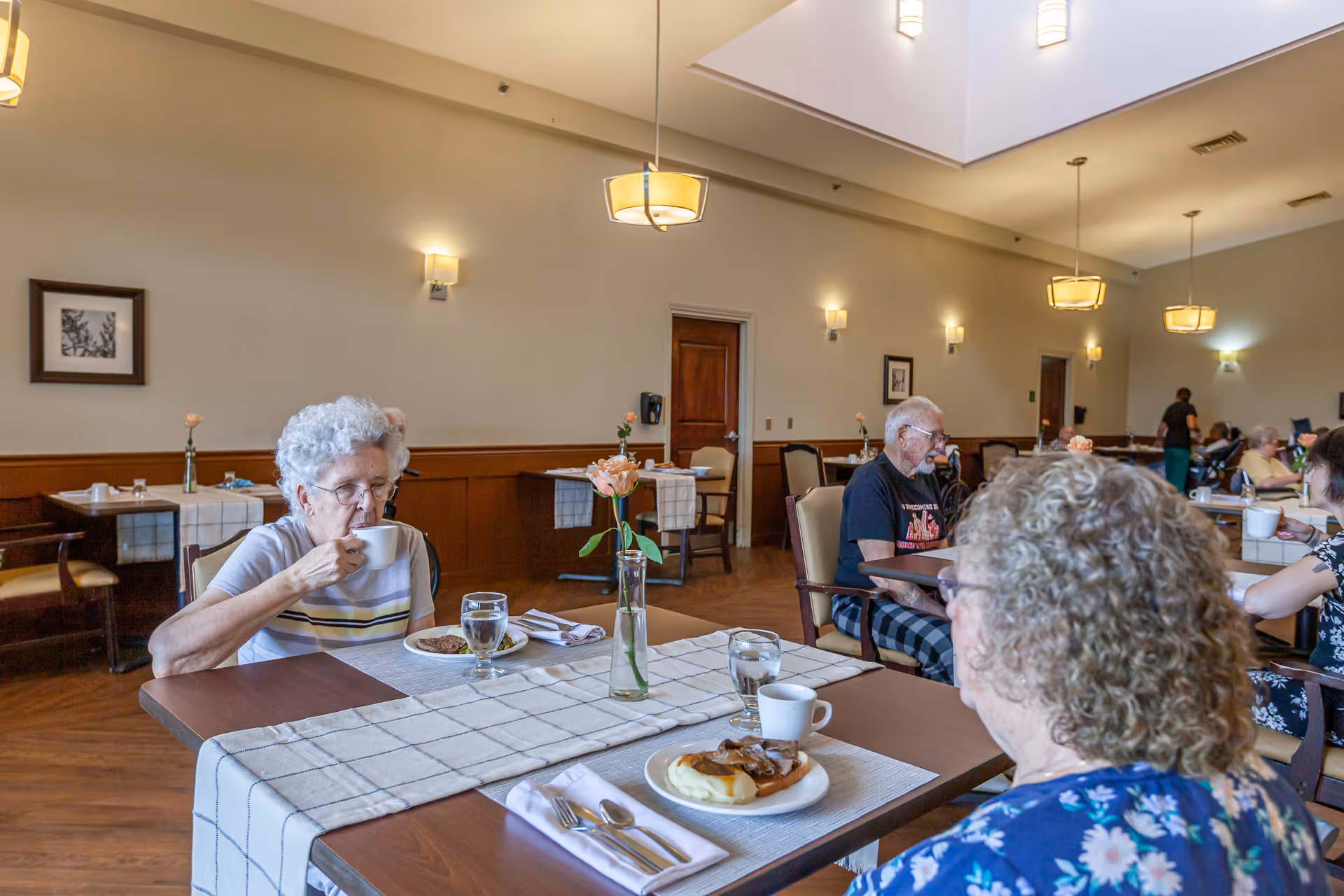 Elderly residents sitting at tables in a well-lit dining room with wooden floors and beige walls, enjoying meals and drinks. The room has hanging pendant lights, framed pictures on the walls, and tables set with checkered tablecloths and flowers in vases.