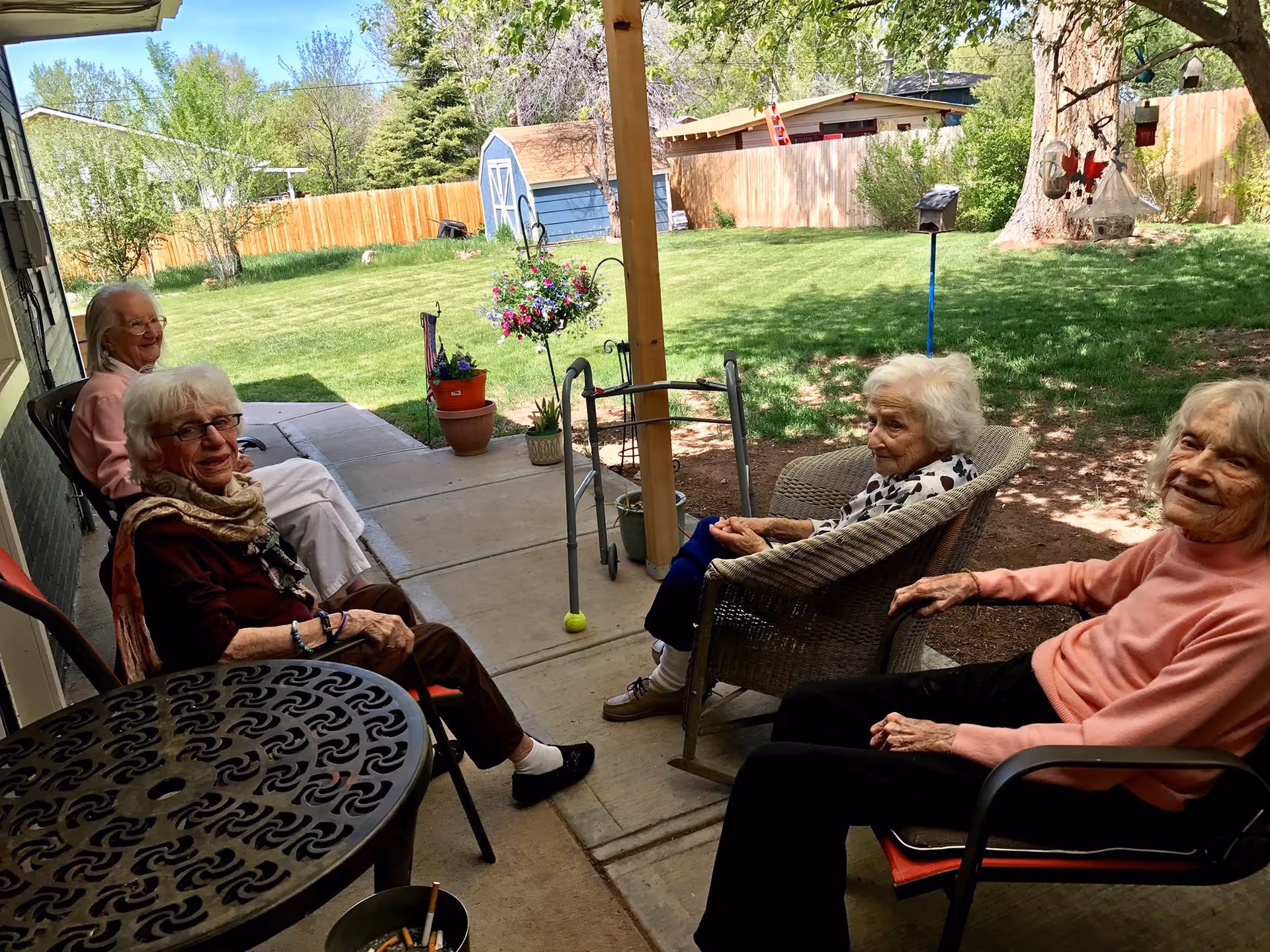 Four elderly women sitting and relaxing on chairs on a covered patio overlooking a green backyard with a wooden fence, a blue shed, and hanging flower pots. A walker is placed near one of the women.