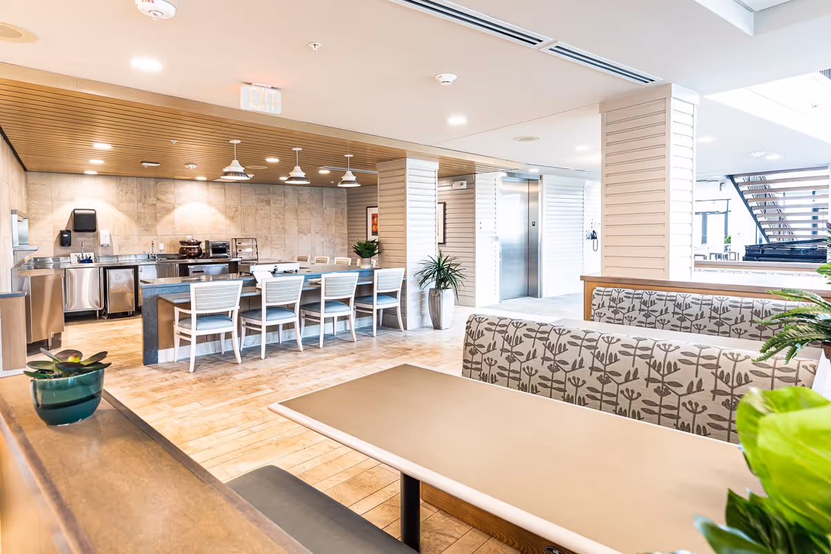 Bright and modern communal dining area with booth seating in the foreground and a long table with white chairs in the background. The space features a kitchen area with stainless steel appliances and a wooden ceiling with pendant lights. There are plants placed around the room and an elevator visible in the background.