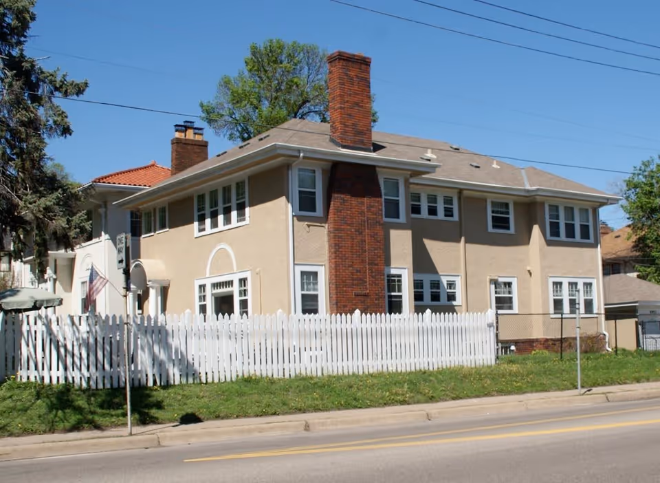 Exterior view of a two-story beige building with multiple windows, a prominent brick chimney, and a white picket fence in front. The building is situated along a street with a sidewalk and some greenery. Trees and a clear blue sky are visible in the background.