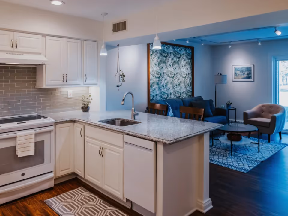 An open kitchen with white cabinets and a granite island overlooking a living room with a sofa, armchair, and artwork.