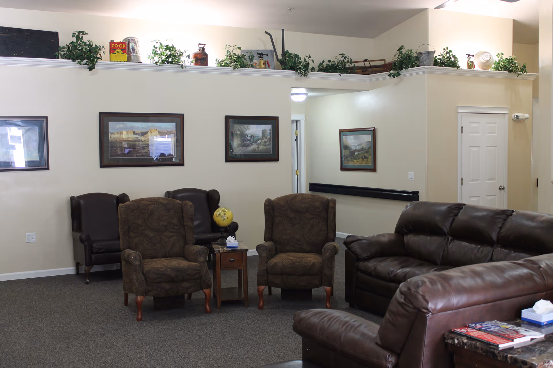 A living room area with two patterned armchairs and two dark leather sofas arranged around a small wooden side table holding a globe and a tissue box. The walls are decorated with framed landscape paintings, and there are green plants and decorative items on a ledge near the ceiling. A white door and a hallway are visible in the background.