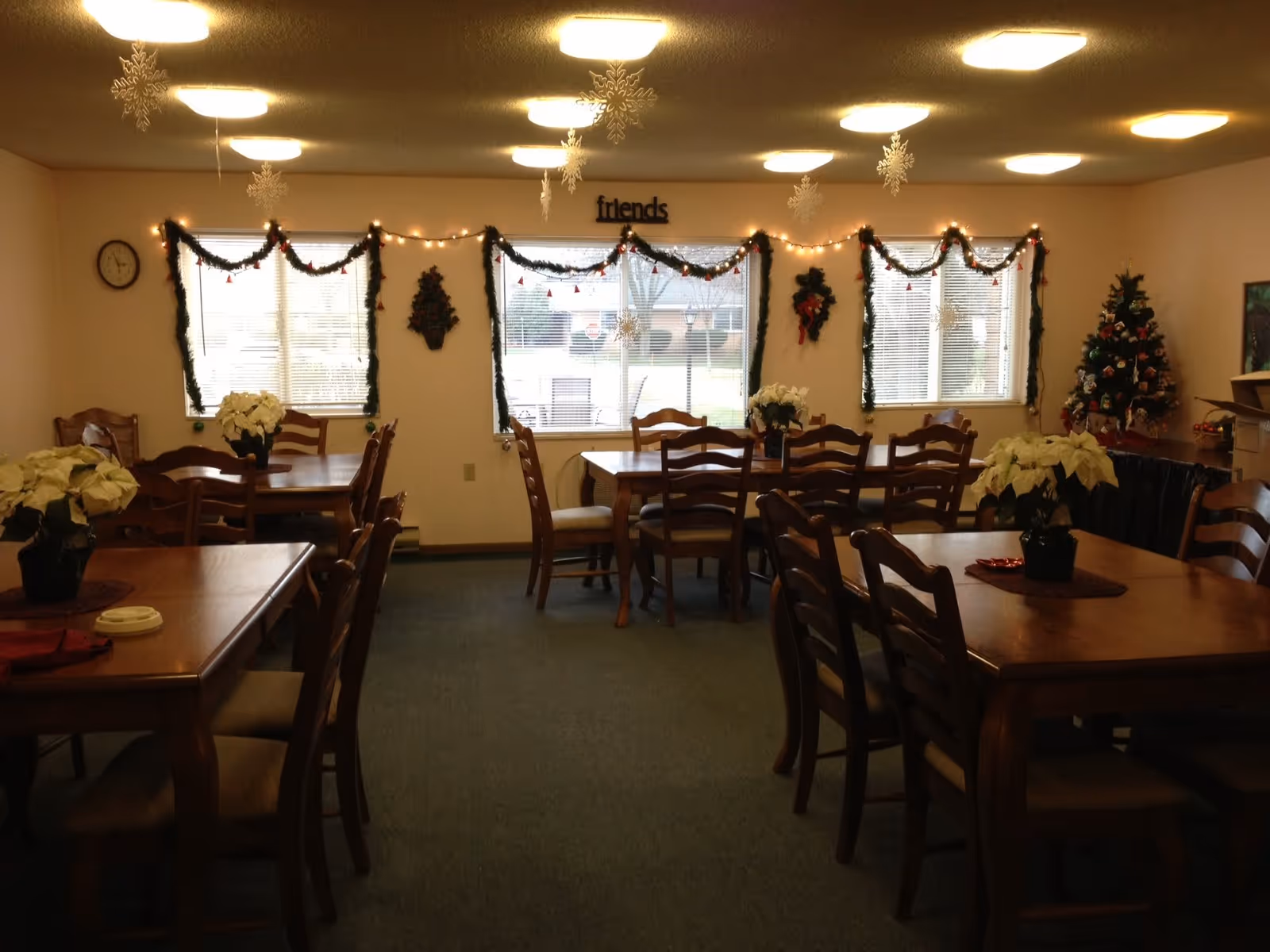 A dining room decorated for the holidays with wooden tables and chairs, white poinsettia plants on the tables, garlands with lights and red ornaments hanging on the windows, snowflake decorations hanging from the ceiling, a Christmas tree in the corner, and a wall clock on the left wall.