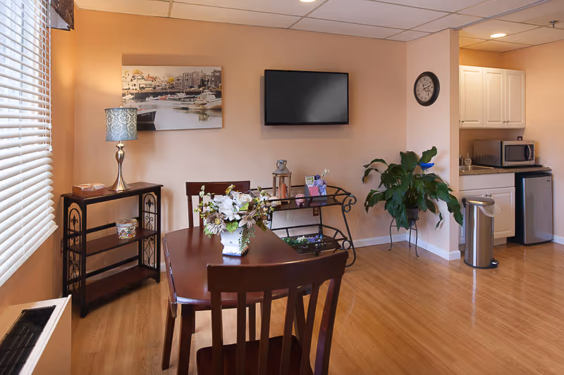 Interior view of a common area in a senior living facility featuring a wooden dining table with chairs, a flower arrangement centerpiece, a small shelving unit with a lamp, a wall-mounted flat screen TV, a clock on the wall, a potted plant, and a kitchenette area with a microwave, mini fridge, and trash can.