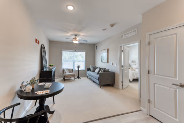 A bright living room with beige walls and carpet. The room features a gray sofa with cushions, a single armchair, a small round table with two chairs, a ceiling fan with light, and a window with blinds letting in natural light. There is a doorway leading to a bedroom with a visible bed and nightstand.