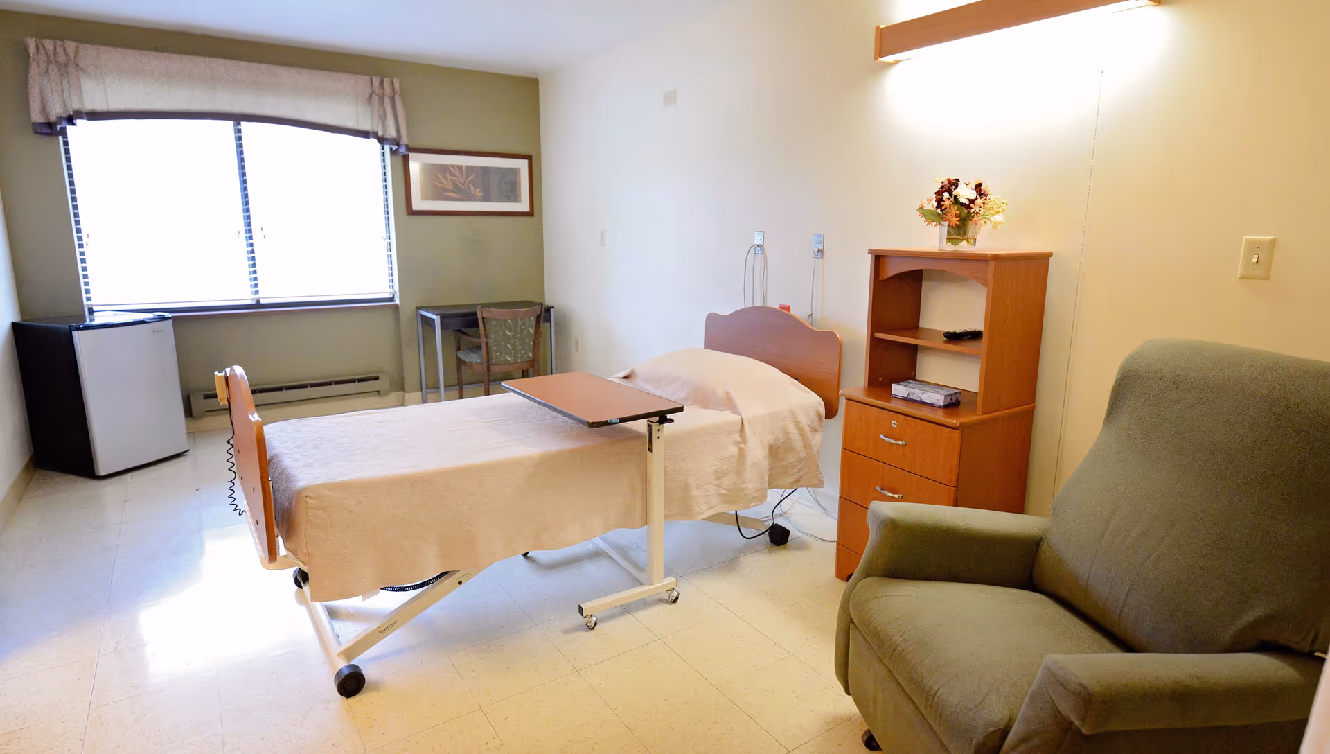A simple, clean room with a single hospital-style bed covered with a beige blanket, a small overbed table, a green upholstered armchair, a wooden cabinet with shelves and drawers, a small table with a chair near a window with blinds and a valance, and a small refrigerator in the corner.