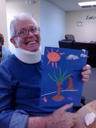 An elderly man wearing glasses and a neck brace is smiling and holding up a colorful craft artwork depicting a sun, a tree, a dog, and a fish. He is seated at a table in a room with chairs and a light-colored wall in the background.