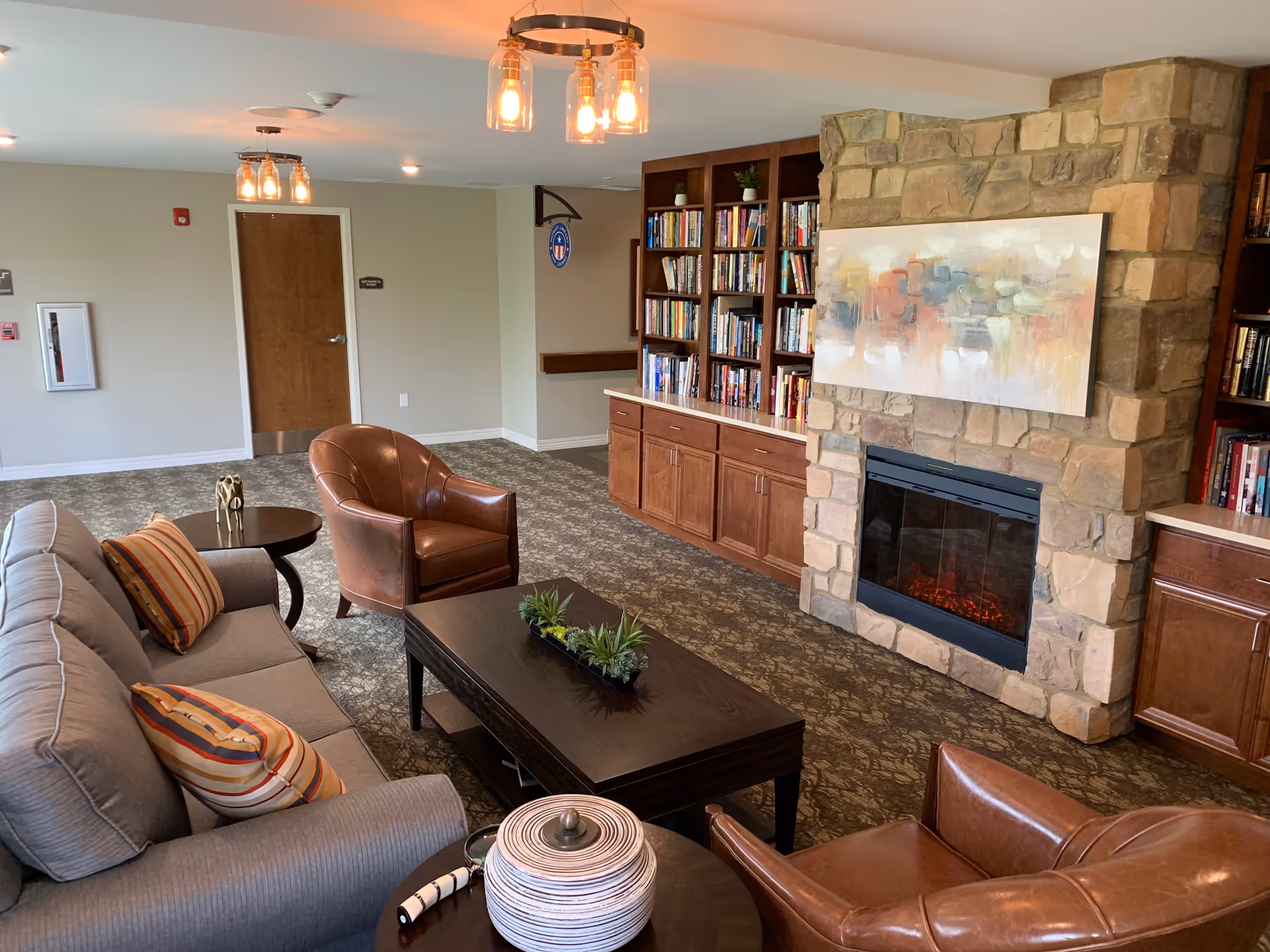 A cozy senior living common area featuring a gray sofa with striped pillows, two brown leather armchairs, a dark wooden coffee table with a small plant centerpiece, and a stone fireplace with an abstract painting above it. Built-in wooden bookshelves filled with books flank the fireplace. The room has patterned carpet and warm lighting fixtures on the ceiling.