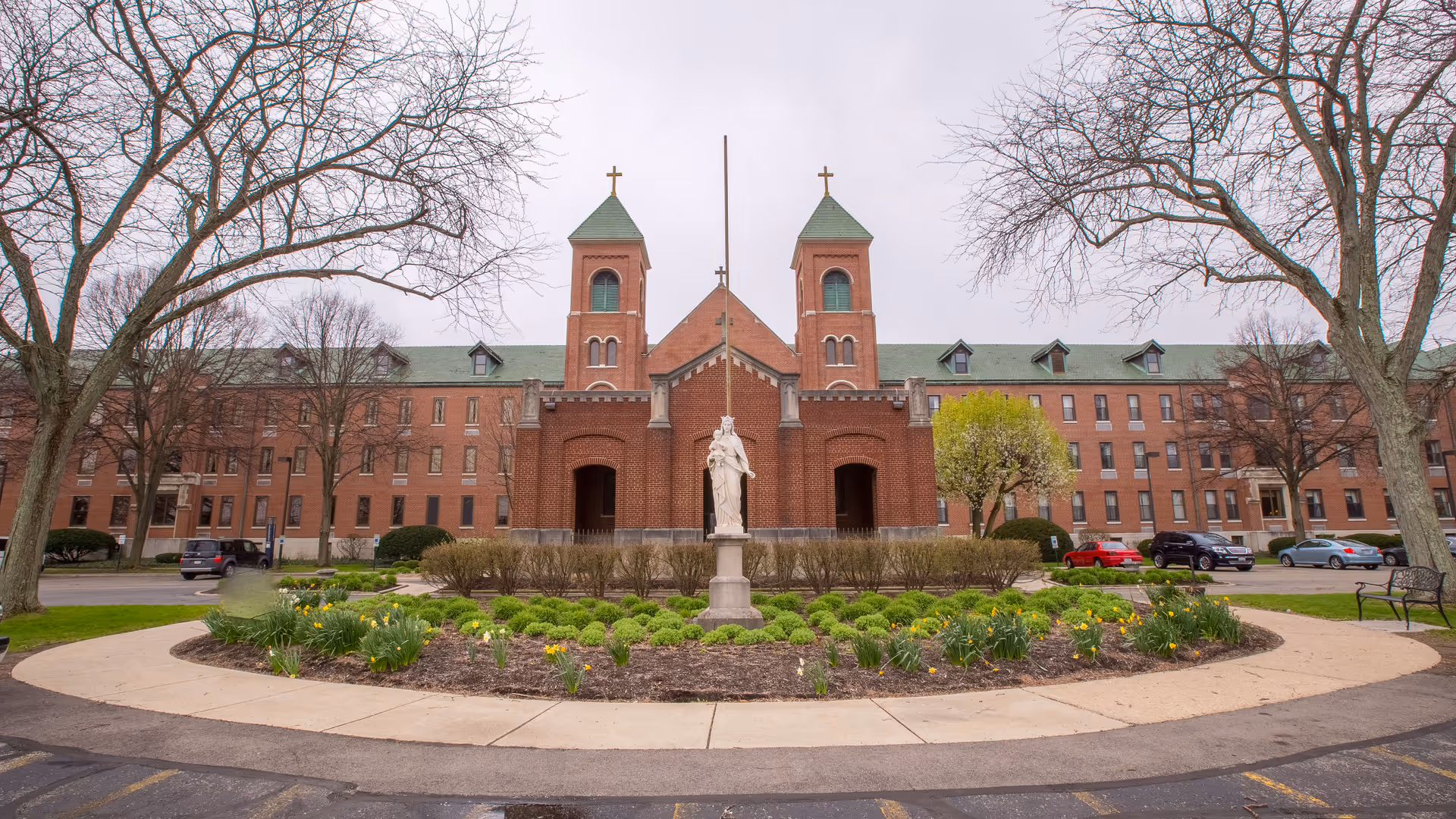 Front exterior view of Maria Joseph Nursing & Rehabilitation Center, a large brick building with two towers topped with crosses. In front of the building is a circular garden with a statue in the center, surrounded by bushes and flowers. Leafless trees frame the scene on both sides, and several cars are parked along the driveway.