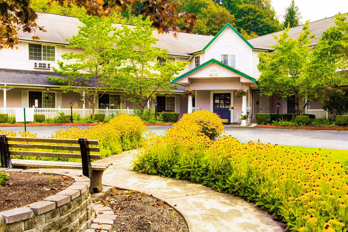 Front exterior view of Cascade Valley Senior Living building with a pathway leading to the entrance, surrounded by lush green trees and yellow flowering bushes, and a wooden bench in the foreground.