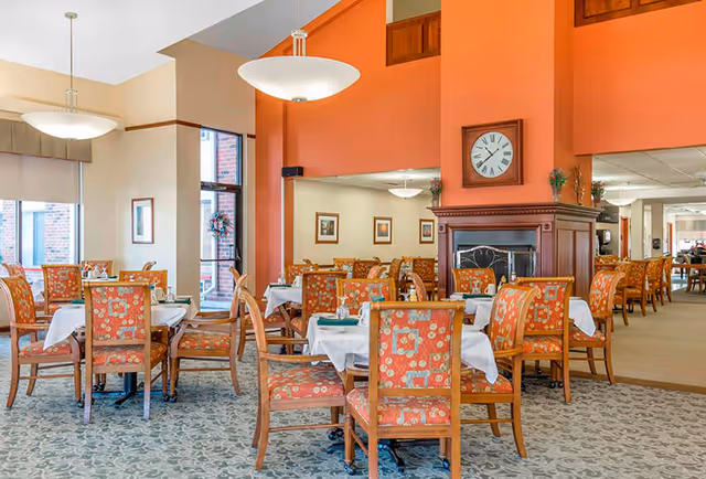 A bright dining room with multiple wooden tables and chairs featuring orange floral upholstery. Each table is covered with a white tablecloth and set with napkins and utensils. The room has large windows letting in natural light, a fireplace with a clock above it, and warm orange and beige walls.