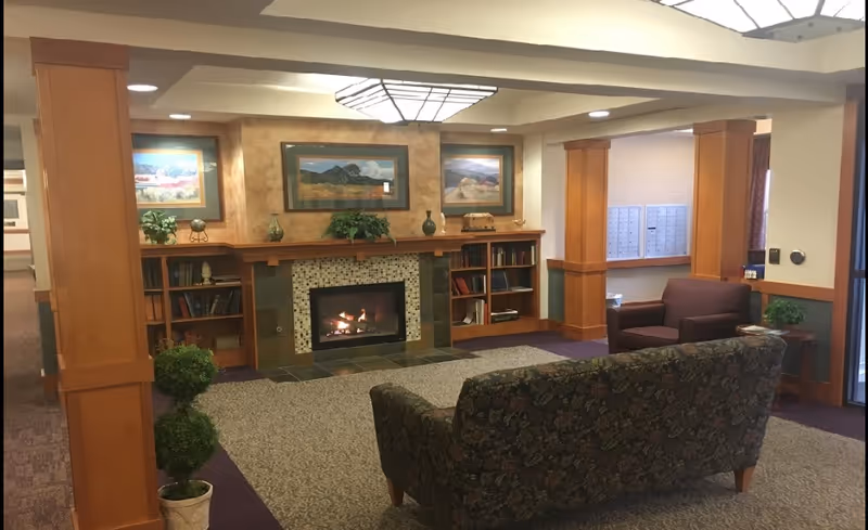 Cozy common room with a lit fireplace, framed artwork, bookshelves, seating, and potted plants.