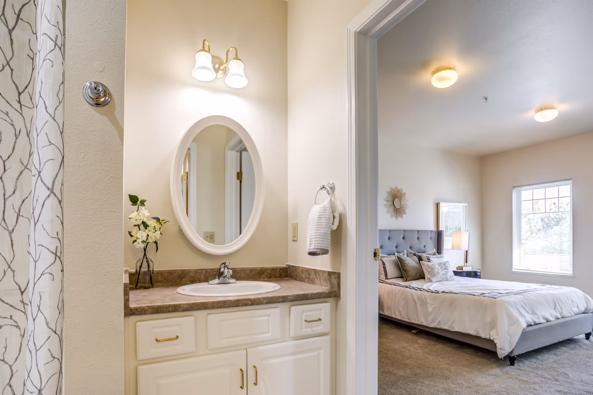 View of a bathroom vanity with a sink, oval mirror, and two light fixtures above. A vase with white flowers is on the countertop. To the left, a shower curtain with a branch pattern is partially visible. Through an open doorway, a bedroom with a neatly made bed, bedside table with a lamp, and a window letting in natural light is seen.
