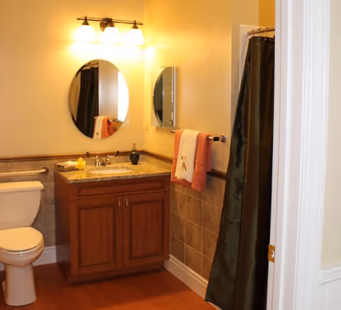 A bathroom with a wooden vanity topped with a granite countertop, an oval mirror above the sink, a toilet to the left, a towel rack with two towels on the right wall, and a shower with a dark curtain partially visible on the right side.