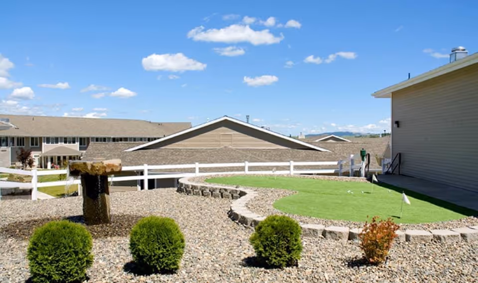 Outdoor area of Bishop Place Senior Living featuring a small putting green with flags, a stone water fountain, neatly trimmed bushes, and a white fence. Residential buildings with beige siding and a clear blue sky with scattered clouds are visible in the background.