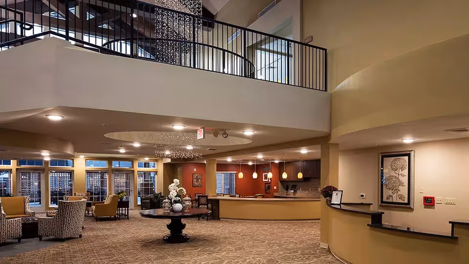 Spacious and well-lit interior common area of The Oxford Grand Assisted Living & Memory Care featuring a large round table with floral arrangements in the center, comfortable seating areas with armchairs, a reception desk, and a mezzanine level with black railings above.
