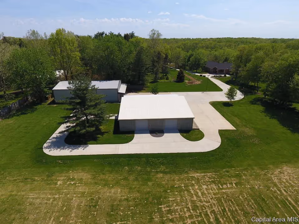 Aerial view of a property with two large white-roofed buildings surrounded by green grass and trees. There is a paved driveway and walkway connecting the buildings, with a forested area in the background under a clear blue sky.