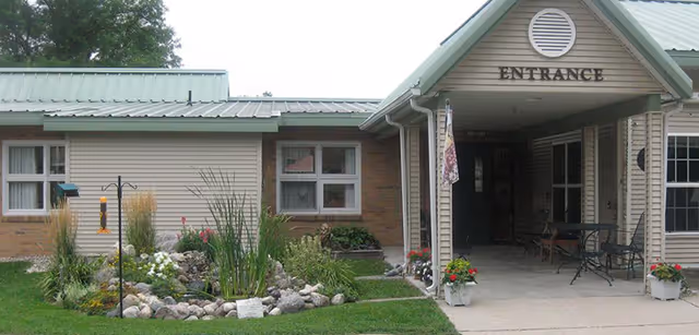 Exterior view of a single-story building with a green metal roof and beige siding. The entrance is covered by a small porch with the word 'ENTRANCE' above it. There are windows on the building and a landscaped garden with rocks, plants, and flowers in front of the building.