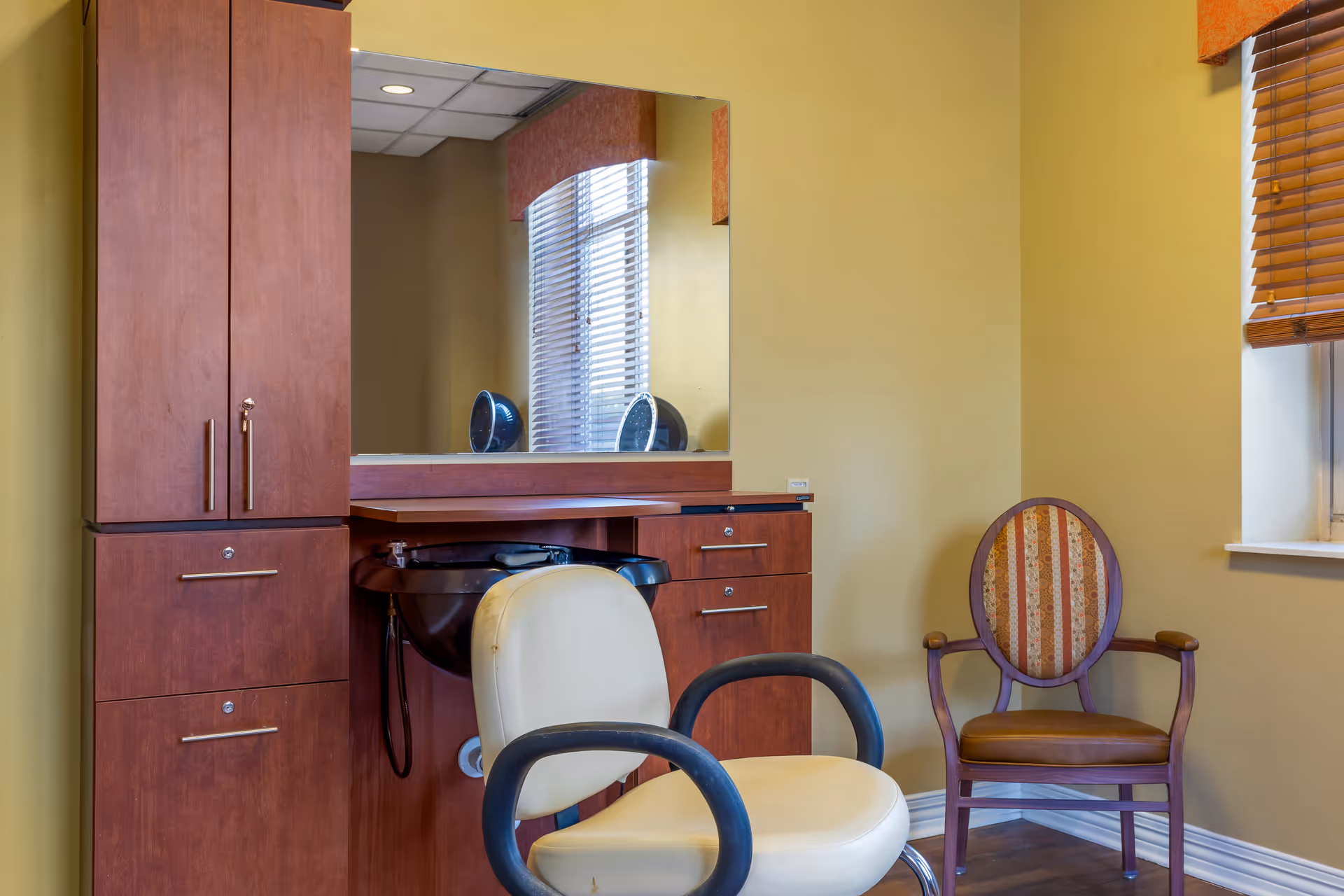 Interior view of a salon area in a senior living facility with a beige salon chair in front of a wooden cabinet and a black wash basin. There is a large mirror above the cabinet reflecting a window with blinds. To the right, there is a wooden armchair with a patterned cushion next to a window with wooden blinds.