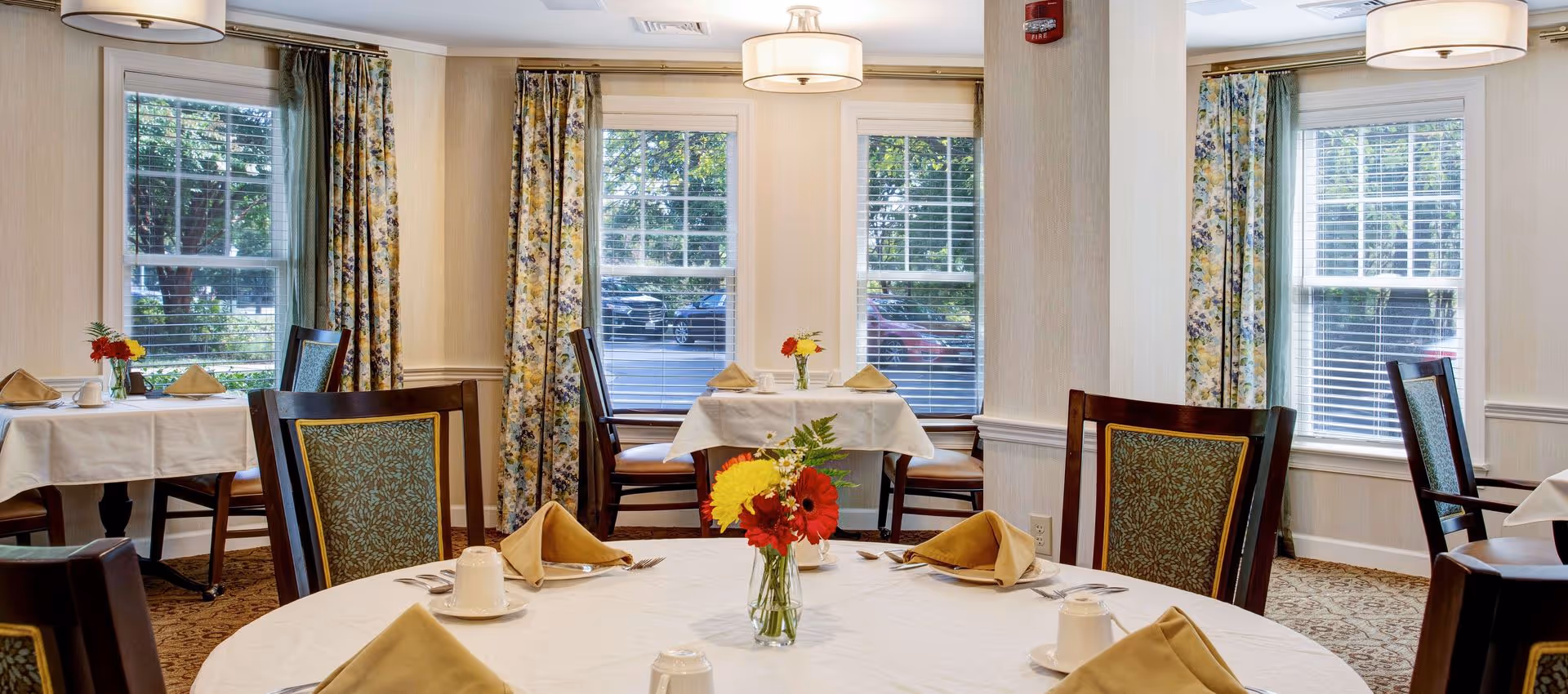 Sunlit dining room with round tables set with napkins and small floral centerpieces, chairs, and large windows with floral curtains.