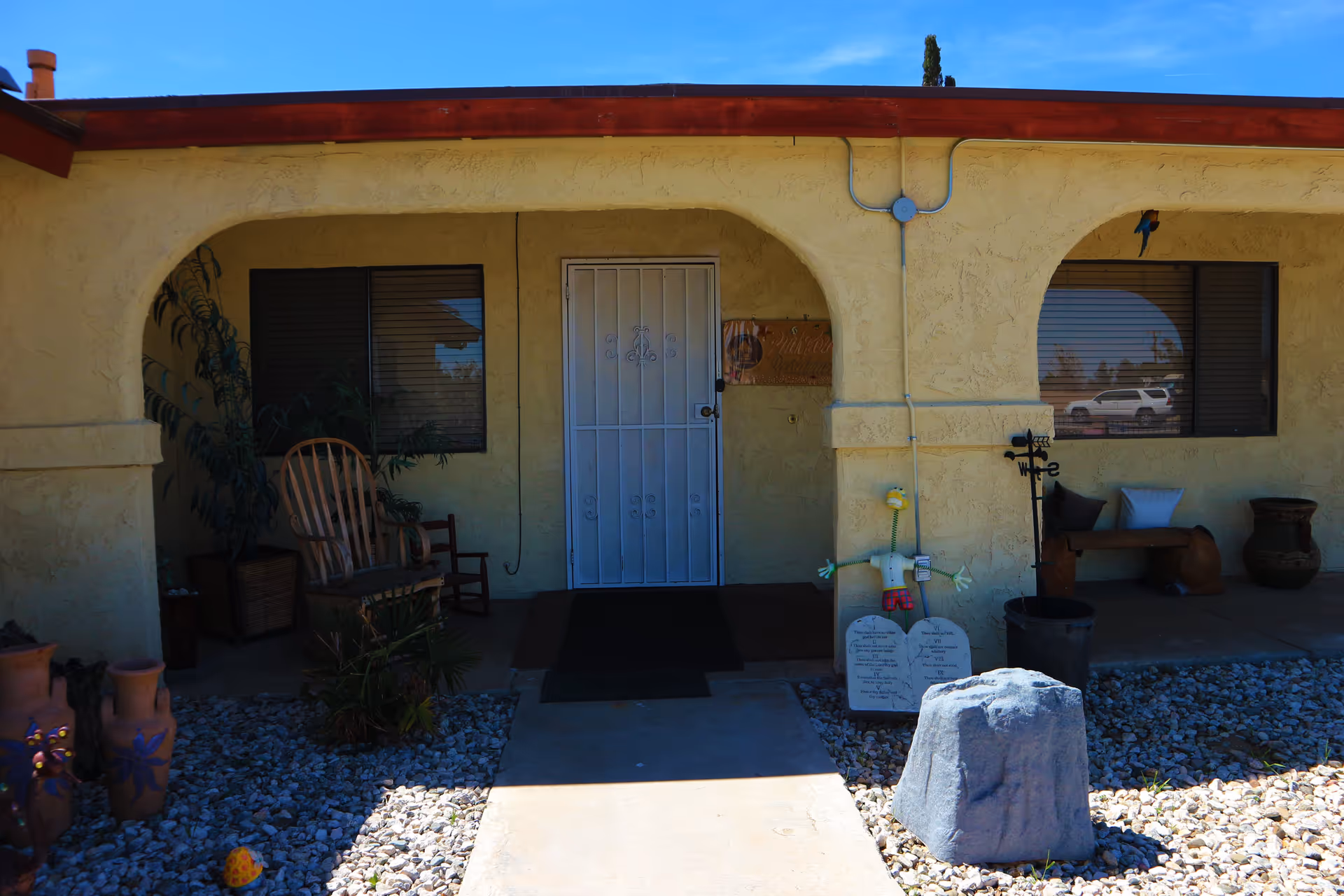 Front entrance of a single-story building with a white metal security door, two windows with blinds, and a covered porch area. The porch has a wooden rocking chair, a small wooden chair, potted plants, decorative items including a figure holding two stone tablets, a weather vane, and a large rock in a gravel yard. The building exterior is textured and painted beige with a reddish-brown trim under a clear blue sky.