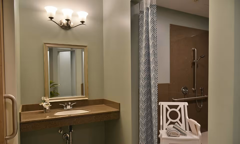 A bathroom with a brown countertop sink, a rectangular mirror above it, and a wall-mounted light fixture with three lamps. To the right, there is a shower area with a grab bar and a handheld showerhead, partially covered by a patterned shower curtain. A white chair with a folded towel is placed near the shower.