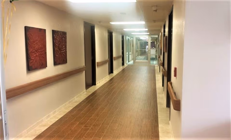 Long, well-lit nursing home hallway with wood-look flooring, handrails along both walls, doors to rooms, and framed wall art.