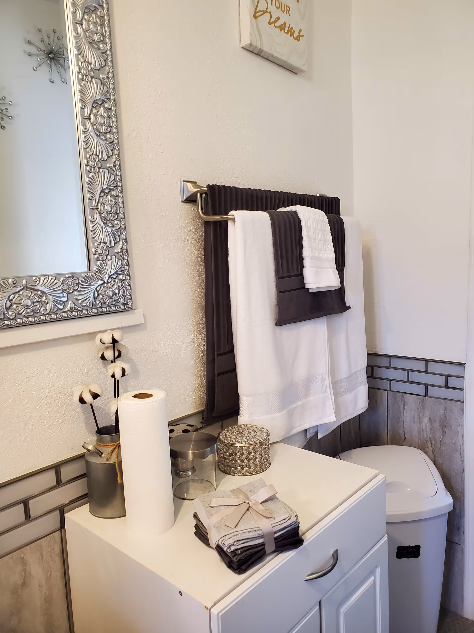 A neatly organized bathroom corner with a white cabinet holding a roll of paper towels, a small metal container with cotton stems, a glass container, a decorative silver container, and a stack of folded towels tied with a ribbon. Above the cabinet, there is a towel rack with dark and white towels hanging. A decorative silver-framed mirror is partially visible on the left wall, and a white trash bin is placed next to the cabinet.