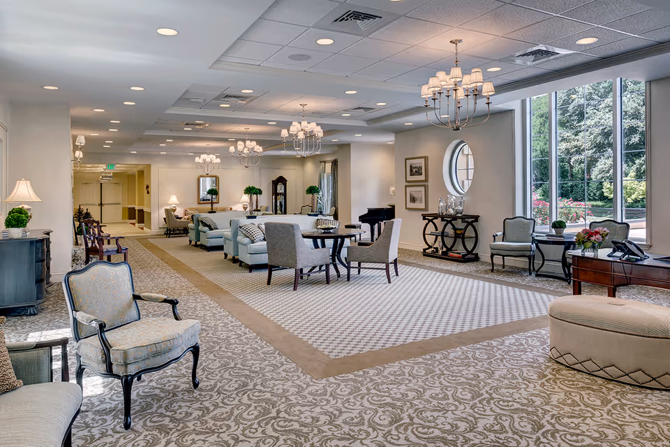 A spacious and well-lit senior living facility common area with patterned carpet, multiple seating arrangements including armchairs, sofas, and tables. The room features large windows allowing natural light, chandeliers hanging from the ceiling, decorative plants, framed artwork, and a piano in the background.