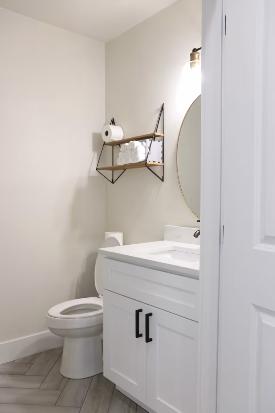 A small bathroom with a white toilet and a white vanity cabinet with a sink. Above the toilet, there is a wall-mounted shelf holding a roll of toilet paper, several rolled white towels, and a small decorative item. A round mirror with a brass frame and a wall light fixture are partially visible above the sink. The floor has a light wood-like tile pattern.