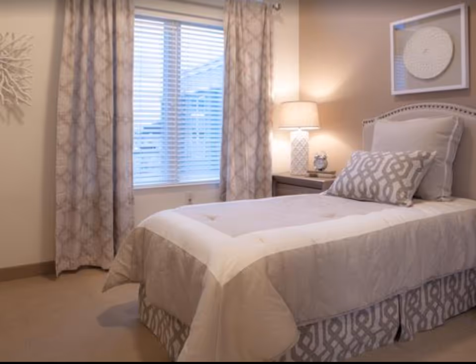 A neatly made single bed in a softly lit bedroom with patterned curtains, a bedside table and lamp, and a window with blinds.