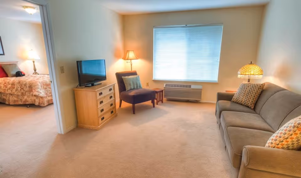 A cozy living room in a senior living facility with a beige carpet, a gray sofa with patterned cushions, a purple armchair with a pillow, a wooden TV stand with a flat-screen TV, two table lamps, and a window with closed blinds. A bedroom with a bed and bedside lamp is visible through an open doorway.