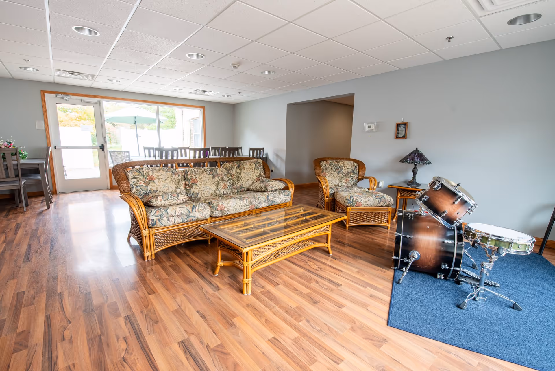 A bright living room with wooden flooring, a floral-patterned wicker sofa and matching armchair, a glass-top wicker coffee table, a small side table with a decorative lamp, and a drum set on a blue rug. In the background, there is a dining area with wooden chairs and a large window with a door leading outside.