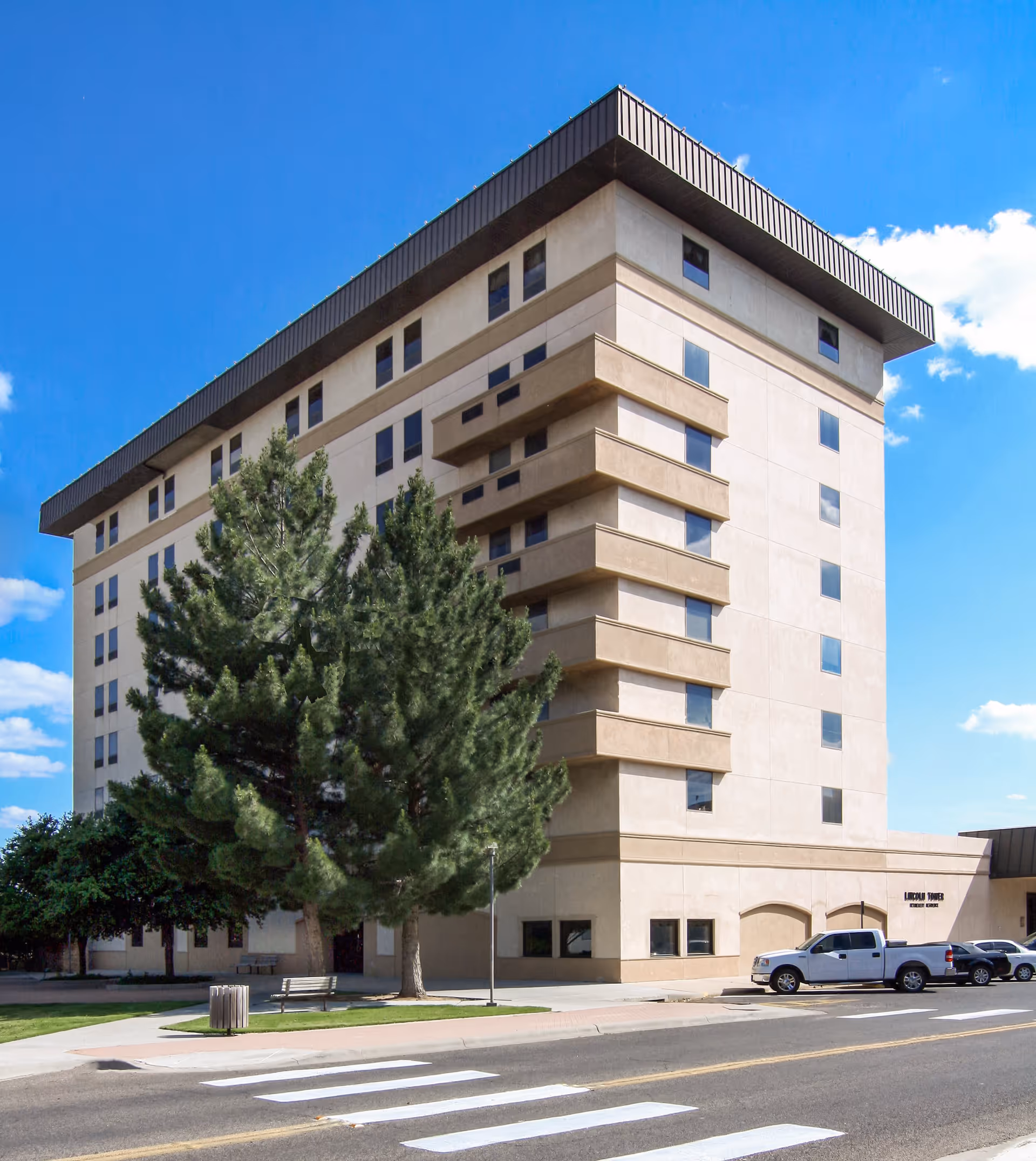 Exterior view of a multi-story beige building named Lincoln Tower Assisted Living with several windows, a few parked vehicles, trees, a bench, and a trash can near the sidewalk under a clear blue sky.