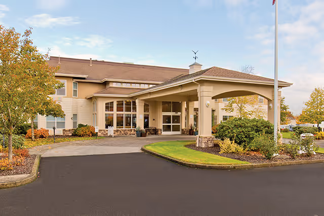 Exterior view of a senior living facility building with a covered entrance, surrounded by landscaped greenery and trees under a partly cloudy sky.