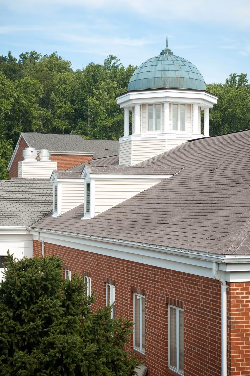 Exterior view of a brick building with a gray shingled roof and a white cupola topped with a green dome. The building is surrounded by green trees under a partly cloudy sky.