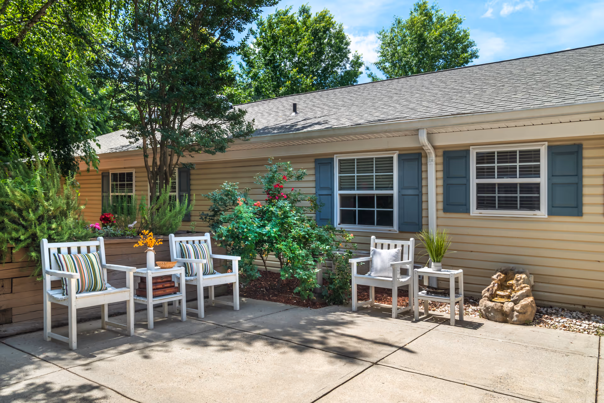 Sunlit outdoor patio with white chairs, small tables, potted plants, and shrubs along the side of a beige building with blue shutters.