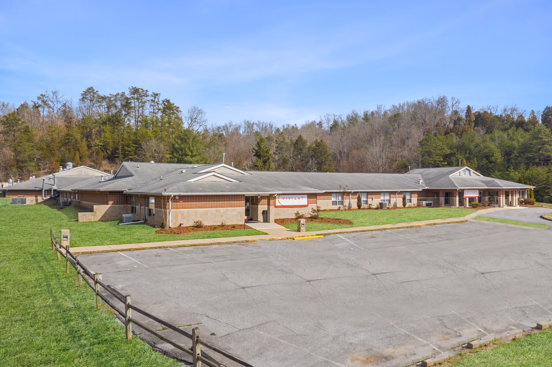 A single-story building with an empty paved parking area, grassy lawns, and wooded hills in the background.