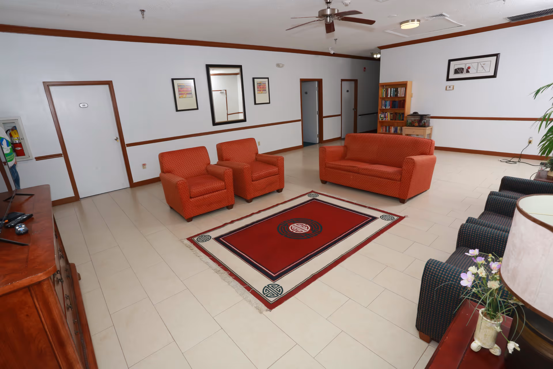 Spacious assisted-living common room with orange armchairs and sofa around a red rug, TV cabinet, bookshelf and additional seating.